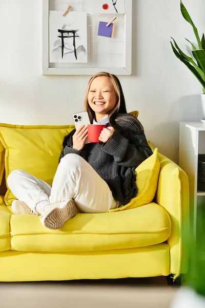Молода азіатка вчиться вдома, сидячи на жовтому дивані, тримаючи чашку. — стокове фото