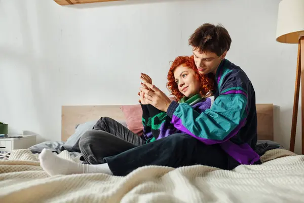 Appealing man with headphones hugging his red haired cheerful girlfriend while on bed at home — Stock Photo
