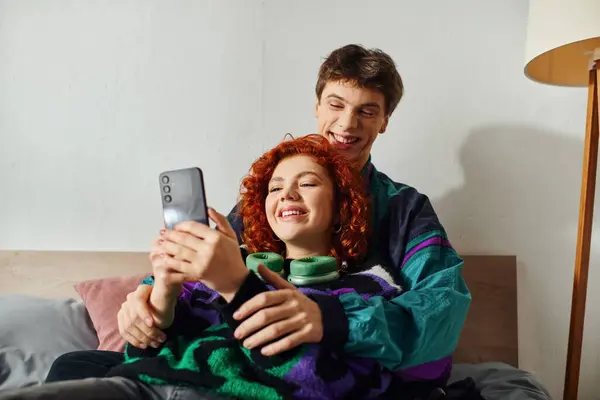 Hombre alegre de buen aspecto con auriculares mirando el teléfono móvil con su novia alegre en la cama - foto de stock