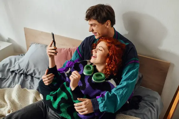 Handsome cheerful man with modern headphones looking at phone with his joyous girlfriend on bed — Stock Photo