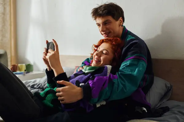 Handsome joyous man with headphones using smartphone with his loving red haired girlfriend on bed — Stock Photo
