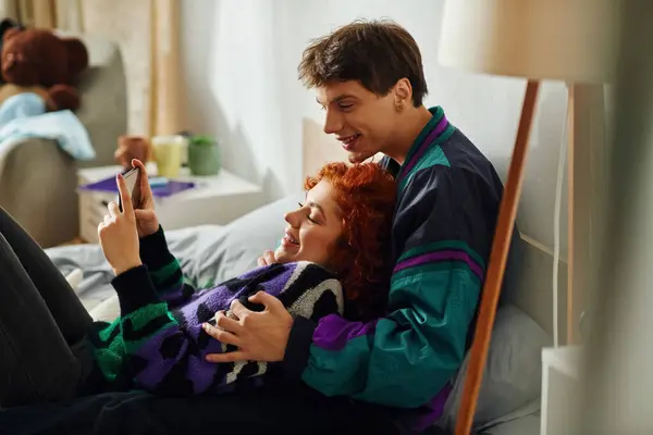 Appealing jolly man with headphones looking at phone with his beautiful cheerful girlfriend — Stock Photo