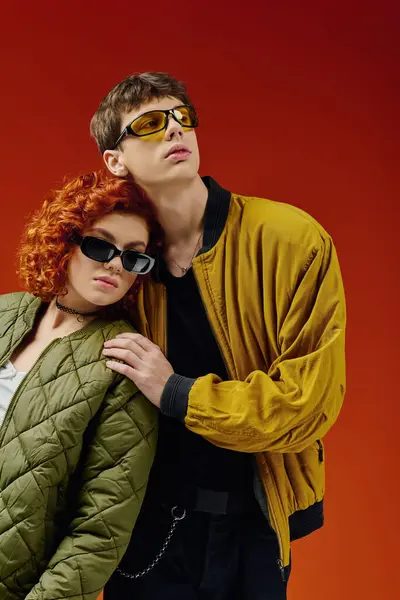 Caring debonair couple in trendy sunglasses in stylish attires posing together on red backdrop — Stock Photo