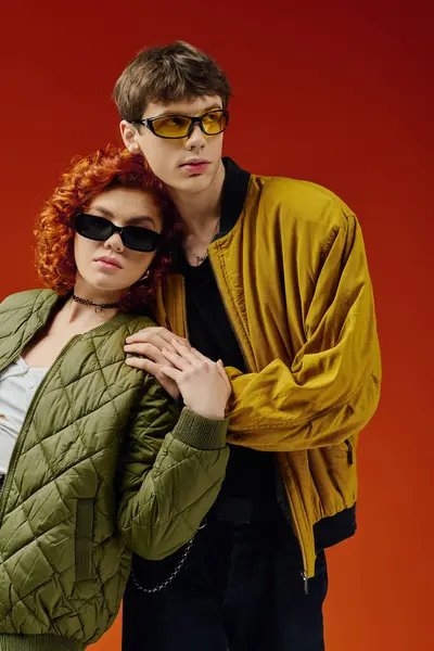 Boa aparência casal amoroso em trajes de moda com óculos de sol posando juntos no fundo vermelho — Fotografia de Stock