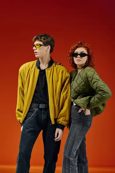 Cool couple attrayant dans des lunettes de soleil à la mode dans des vêtements chics posant ensemble sur fond rouge — Photo de stock