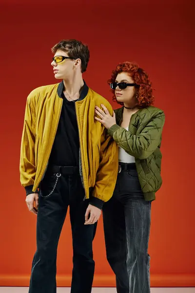 Liebendes Debonair-Paar in schicker Sonnenbrille in trendigen Anzügen posiert gemeinsam vor rotem Hintergrund — Stockfoto
