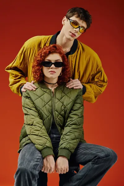 Приваблива пара в стильних вбраннях з сонцезахисними окулярами, що сидять і позують разом на червоному тлі — стокове фото