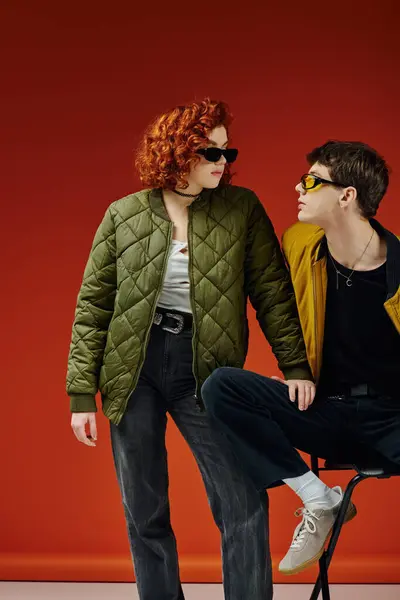 Good looking man with sunglasses sitting on chair near his red haired girlfriend on red backdrop — Stock Photo