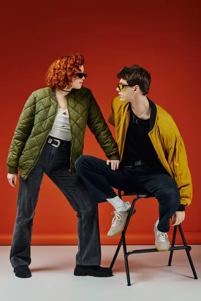 Young man in fashionable attire with sunglasses sitting on chair next to his red haired girlfriend — Stock Photo