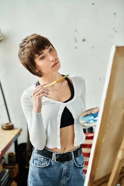 Artistically dressed woman stands before easel, pencil in hand. — Stock Photo