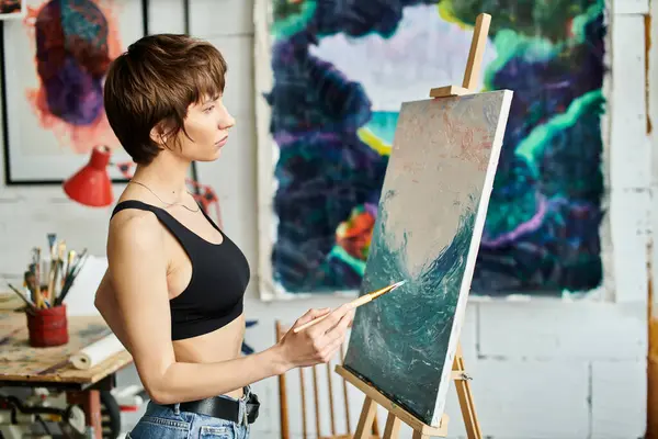 Woman artist standing at easel, holding paintbrush. — Stock Photo