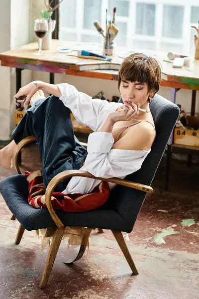 Mujer joven con estilo elegante se sienta en una silla, fumando un cigarrillo en el estudio de arte. - foto de stock