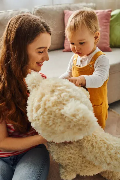 A young mother lovingly holds a stuffed animal next to her toddler daughter at home, creating a heartwarming moment of affection. — Stock Photo