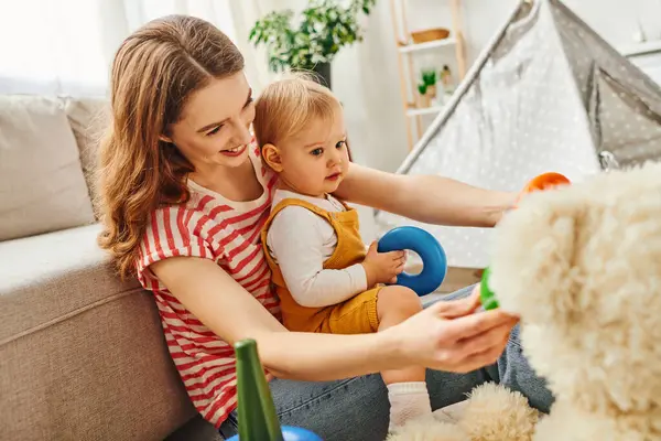 A young mother lovingly cradles her toddler daughter while sitting on a cozy couch at home. — Stock Photo