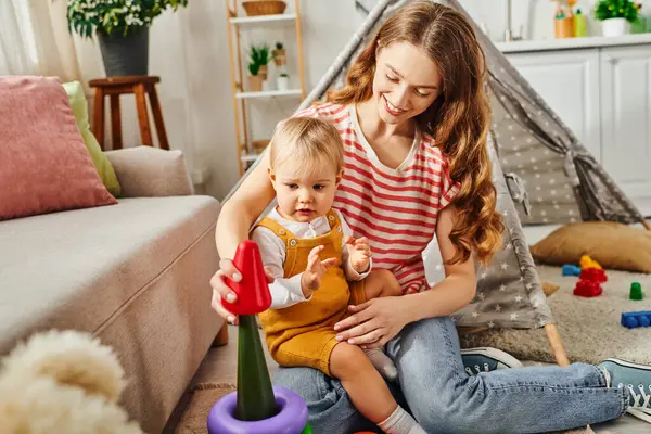 A young mother lovingly holds her baby while engaging in playtime with a colorful toy. — Stock Photo