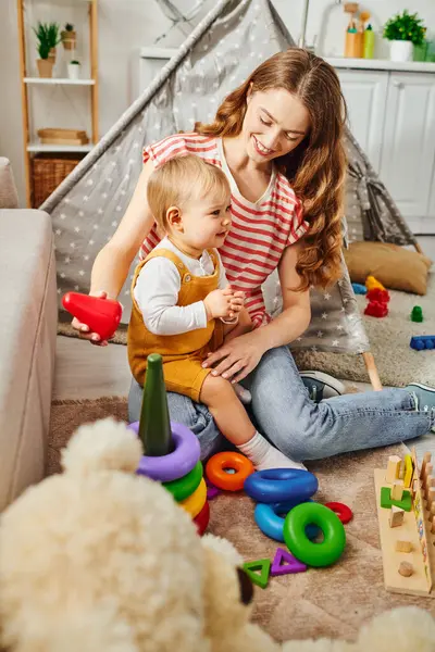 Una joven madre juega alegremente con su hija pequeña en el suelo en casa, uniéndose y creando recuerdos felices juntos. - foto de stock