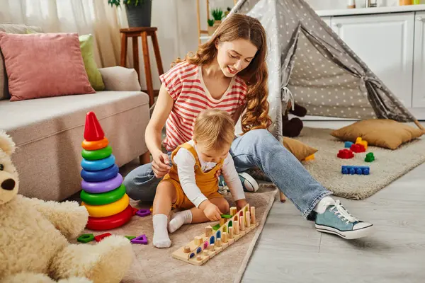 Una giovane madre sta giocando con gioia con la sua bambina sul pavimento di casa, favorendo un forte legame attraverso attività divertenti. — Foto stock
