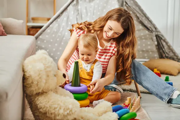 Una madre joven y su hija pequeña jugando felizmente con un oso de peluche, creando recuerdos conmovedores en casa. - foto de stock