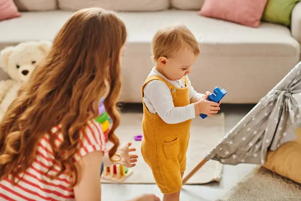 Ein kleines Kind erkundet in Begleitung seiner Mutter fröhlich das Wohnzimmer mit einem bunten Spielzeug. — Stockfoto