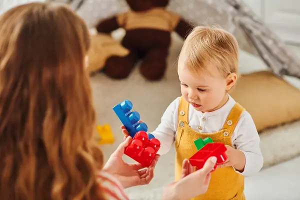 Una madre joven sostiene cariñosamente a su hija bebé mientras juegan alegremente con juguetes coloridos juntos en casa. - foto de stock