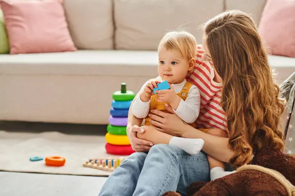 A young mother lovingly holds her toddler daughter while sitting on a cozy couch at home. — Stock Photo
