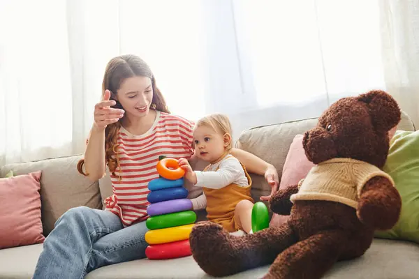 Uma jovem mãe se senta em um sofá com sua filha bebê e um ursinho de pelúcia, compartilhando um momento terno de amor e união. — Fotografia de Stock