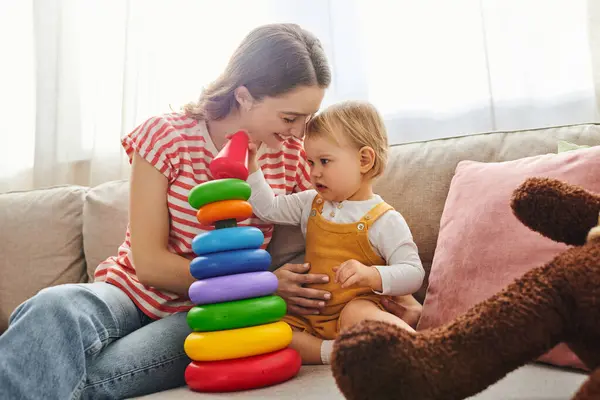 A young mother and her toddler daughter laughing and playing together on a couch in a cozy home setting. — Stock Photo