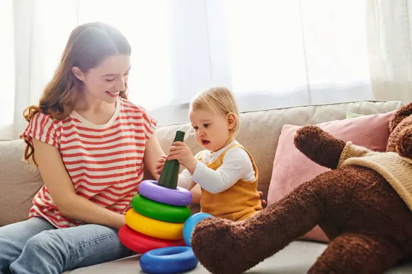 A young mother sits on a cozy couch, tenderly playing with her smiling toddler daughter in a heartwarming moment of love. — Stock Photo