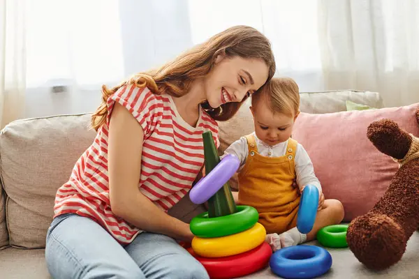 A young mother and her toddler daughter laughing and playing on a cozy couch, creating precious memories together. — Stock Photo