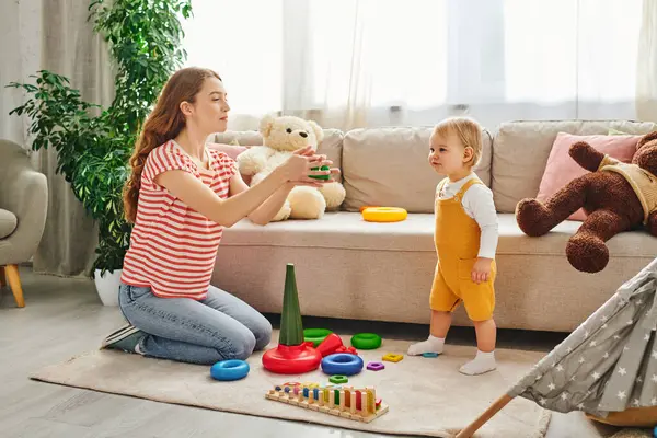 Una joven madre se involucra juguetonamente con su hija en medio de juguetes en una acogedora sala de estar. - foto de stock