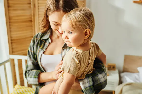Une jeune mère tient amoureusement sa petite fille dans ses bras à la maison, créant un moment de connexion chaleureux. — Photo de stock