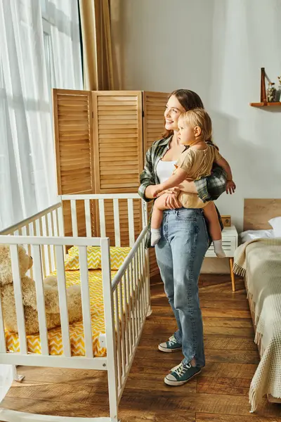 A young mother lovingly cradles her baby daughter in a white crib, creating a heartwarming scene of maternal care and bonding. — Stock Photo