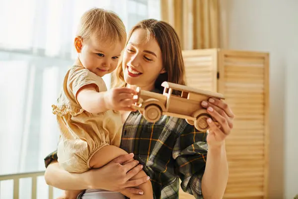 Uma jovem mãe segura sua menina enquanto eles brincam com um carro de brinquedo, colagem e criação de memórias estimadas em casa. — Fotografia de Stock