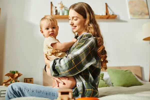 A young mother sitting on a bed, gently holding her baby daughter in her arms, cherishing a precious moment together. — Stock Photo