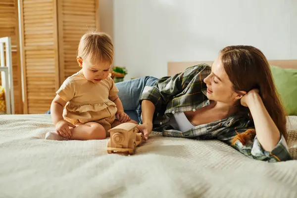 Uma jovem mãe deita-se em uma cama com sua filha bebê, ambos sorrindo, enquanto um brinquedo descansa ao lado deles. — Fotografia de Stock