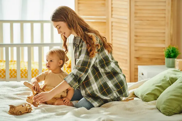 Junge Mutter sitzt auf einem Bett neben ihrer kleinen Tochter und teilt einen Moment der Liebe und Verbundenheit in einer gemütlichen häuslichen Umgebung. — Stockfoto