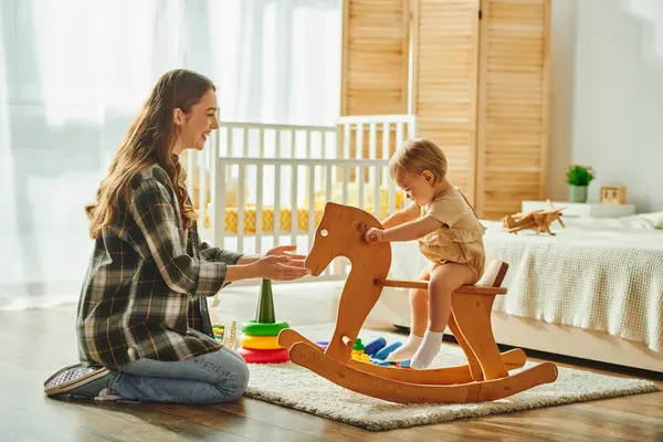 Una giovane madre gioca felicemente con la sua bambina su un cavallo a dondolo in legno, creando dolci ricordi a casa.. — Foto stock