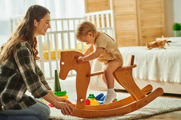 Uma menina jovialmente brinca com um cavalo de balanço de madeira enquanto sua mãe assiste e sorri em sua casa acolhedora. — Fotografia de Stock