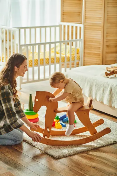 Uma jovem mãe brinca alegremente com sua filha criança em um cavalo de balanço de madeira tradicional, colagem e criação de memórias estimadas. — Fotografia de Stock