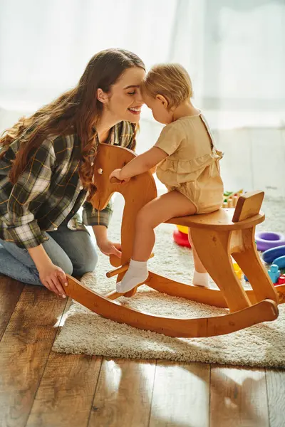 A young mother joyfully playing with her toddler daughter on a rocking horse at home. — Stock Photo