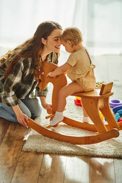 Uma jovem mãe brincando alegremente com sua filha criança em um cavalo de balanço em casa. — Fotografia de Stock