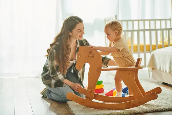 Una joven madre juega alegremente con su hija pequeña en un caballo mecedora de madera en su acogedora casa. - foto de stock