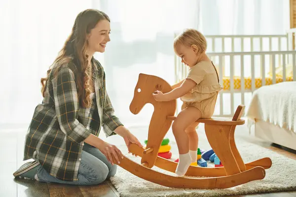 Uma jovem mãe brinca alegremente com sua filha criança em um cavalo de baloiço em casa, unindo e criando memórias especiais. — Fotografia de Stock