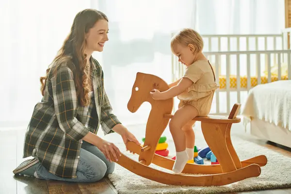 Una joven madre juega felizmente con su hija pequeña en un caballo mecedora en casa, uniéndose y creando recuerdos especiales juntos. — Stock Photo