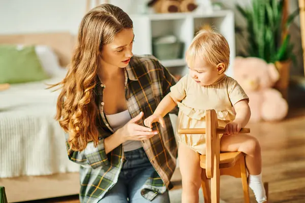 A young mother sitting in a chair beside her toddler daughter, sharing a warm moment together at home. — Stock Photo