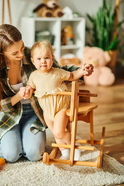 A young mother kneels next to her toddler daughter sitting in a high chair, sharing a precious moment together at home. — Stock Photo