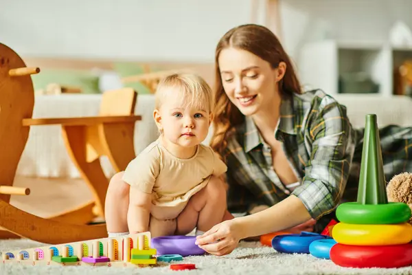 Радісна молода мати взаємодіє зі своєю дочкою малюка на затишній підлозі свого будинку. — стокове фото