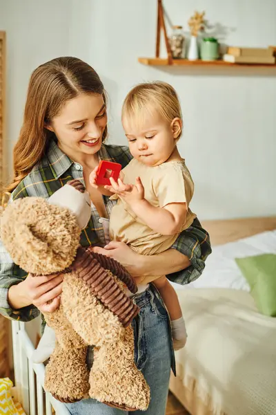 A young mother lovingly holds her baby daughter while they both enjoy a cuddly teddy bear at home. — Stock Photo