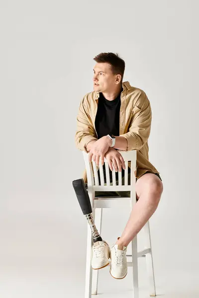 Un hombre guapo con una pierna protésica se sienta confiadamente encima de una silla blanca. - foto de stock
