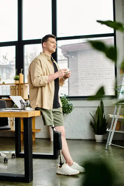 Un hombre de negocios guapo con una pierna protésica de pie frente a una ventana, sosteniendo una taza. - foto de stock